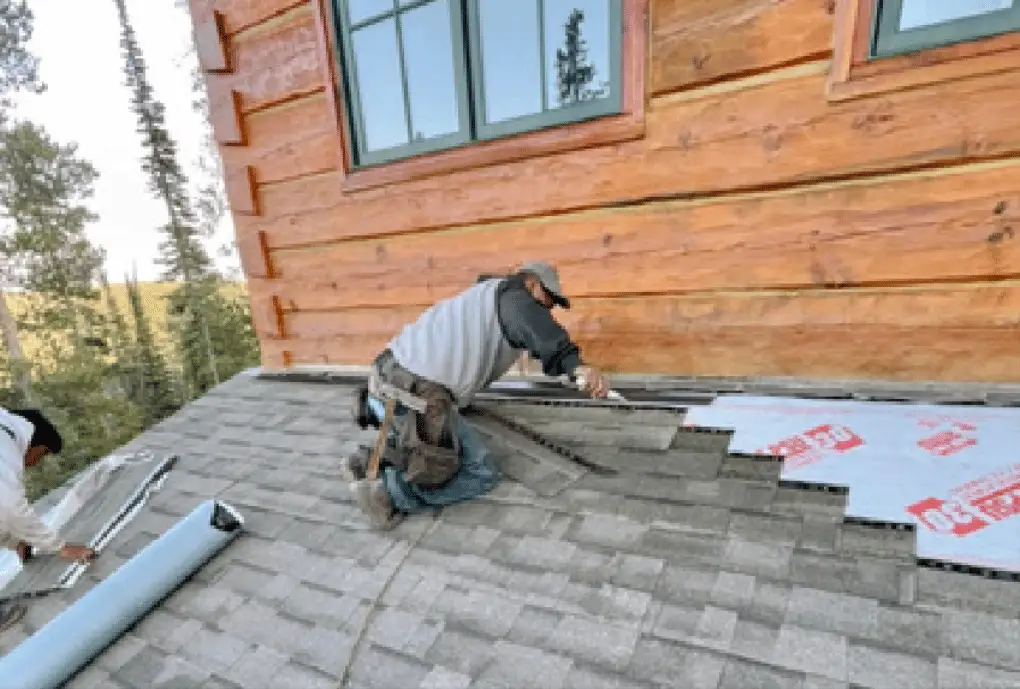 Roofer installing grey shingles over underlayment beside a log wall