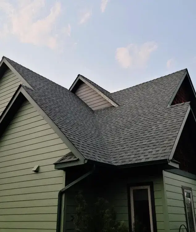 Green-sided home with a new dark grey shingle roof over several gables at dusk