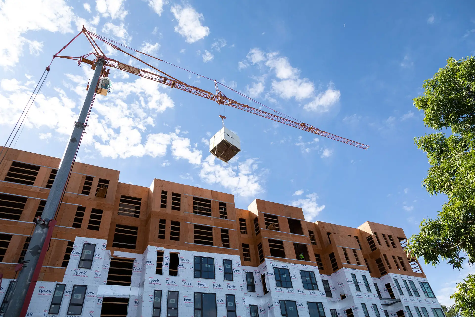 Crane lifting a rooftop unit onto a wood-framed multi-storey building wrapped in house wrap