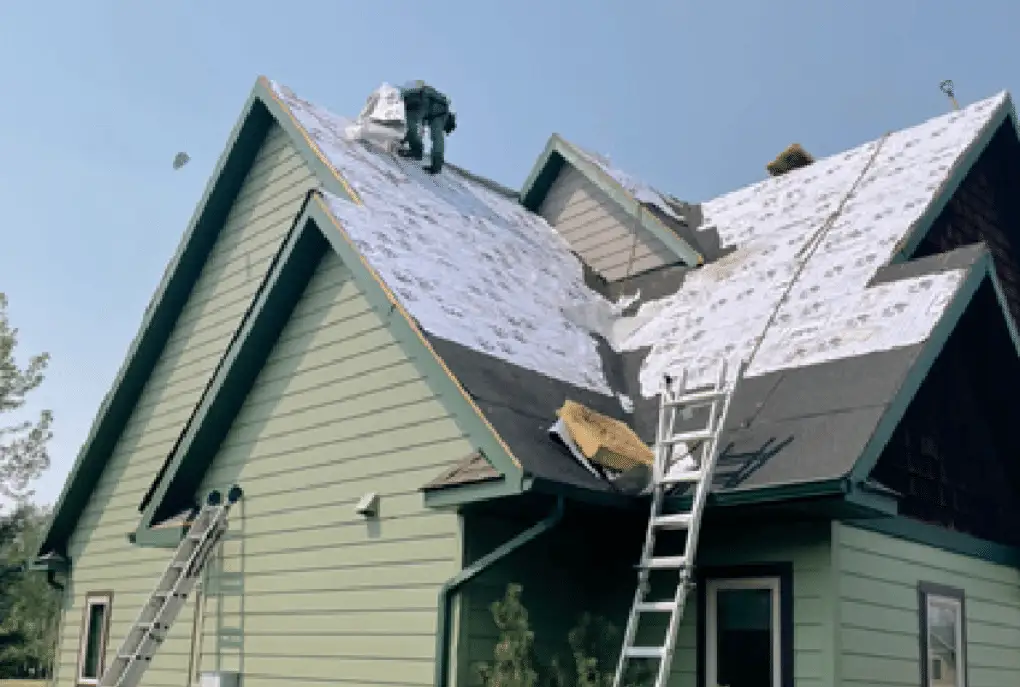 Roofer tearing off an old roof on a green home, underlayment and ladders in place