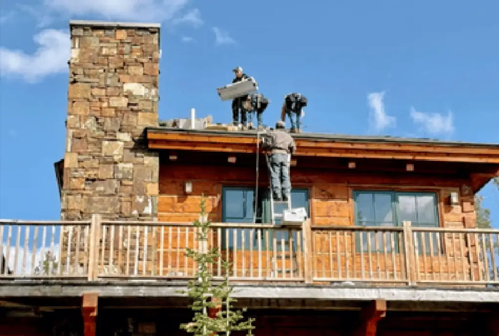 Crew carrying roofing materials onto a log home beside a stone chimney