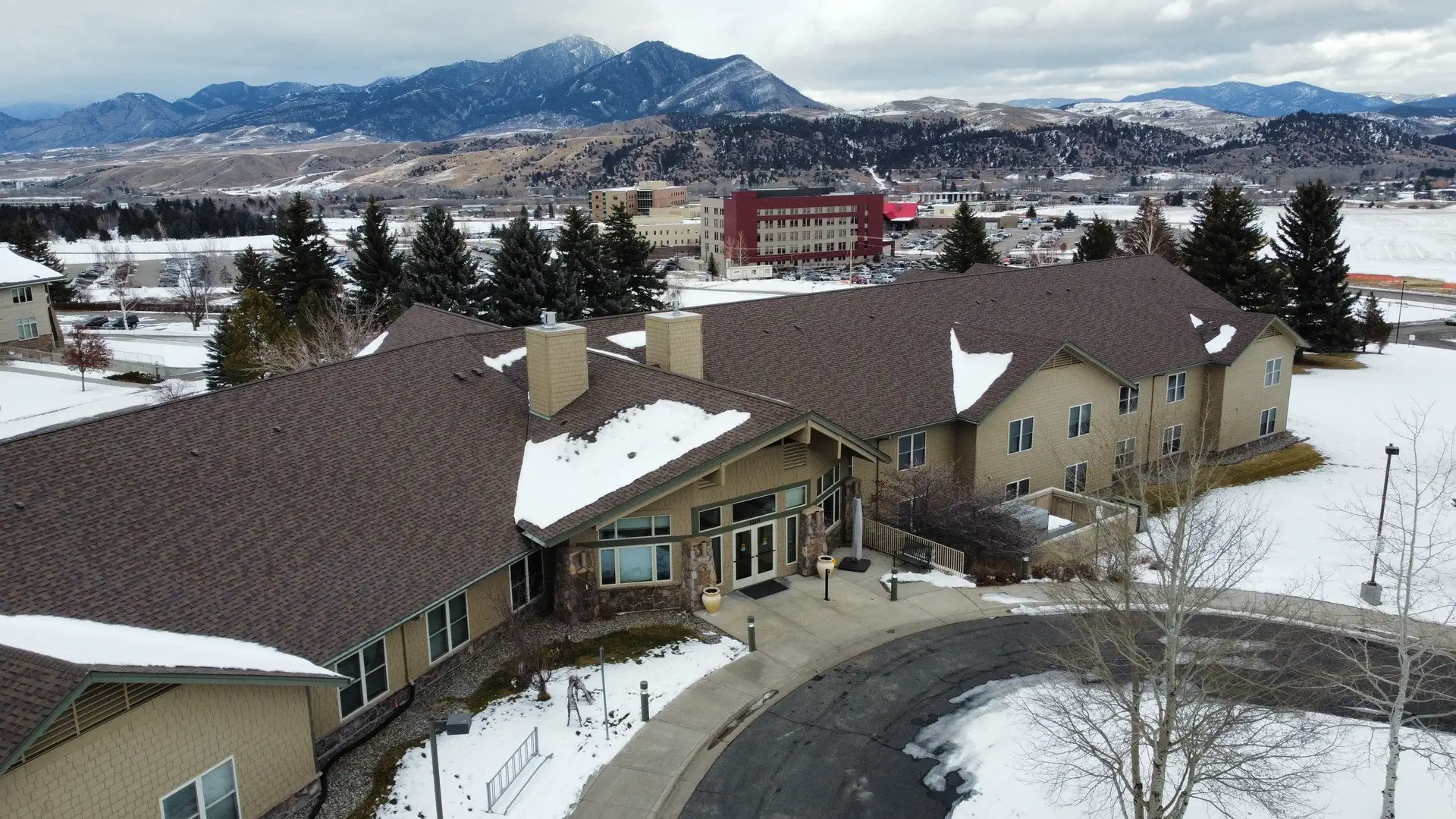 Aerial view of a large building with snow-dusted brown shingle roofs and mountains behind