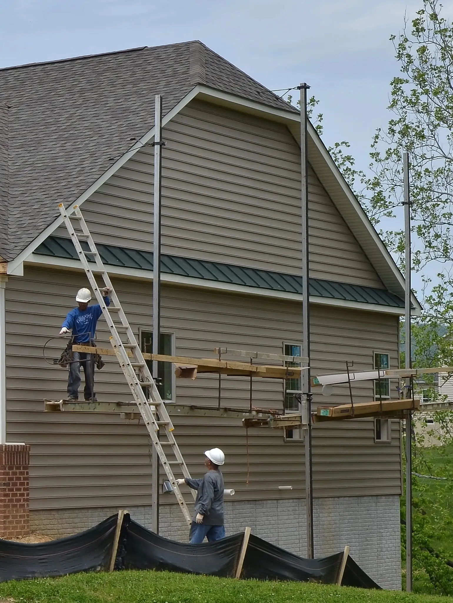 Two workers in hard hats on scaffolding installing tan vinyl siding