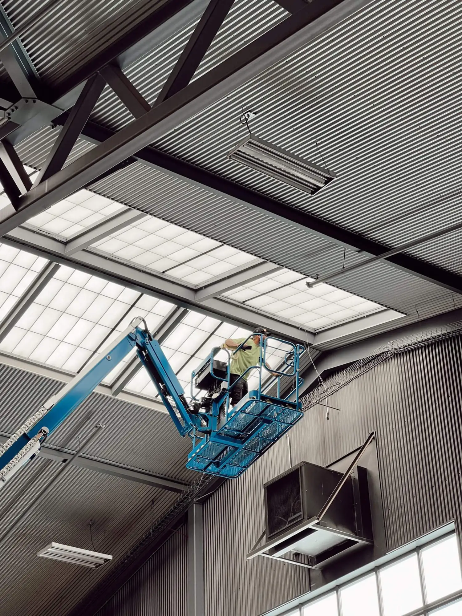 Worker on a boom lift inspecting the metal roof and skylights inside a large building